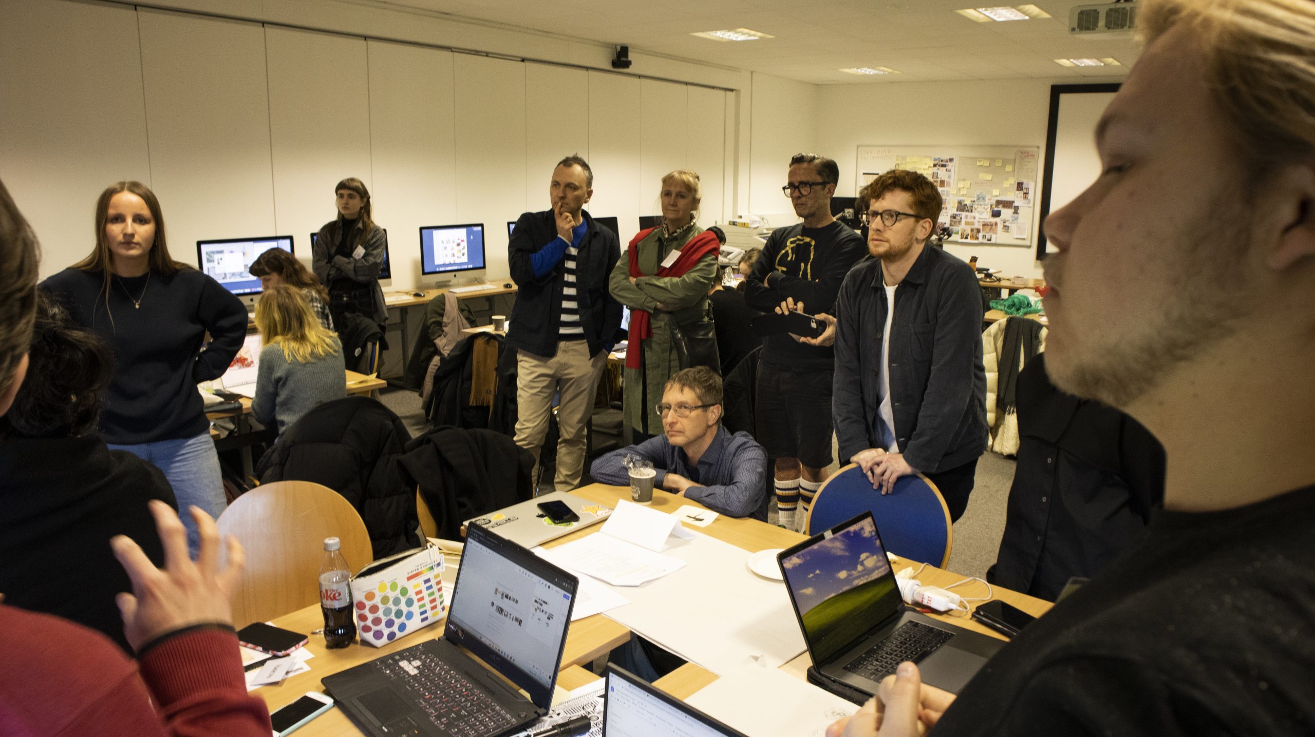 A group of people gathered in a classroom or office-like environment. Several individuals are standing, some with thoughtful expressions, while others are seated at desks with laptops and papers spread out. In the background, computer monitors display various content, and whiteboards or bulletin boards are visible on the walls. The setting suggests a collaborative or brainstorming session.
