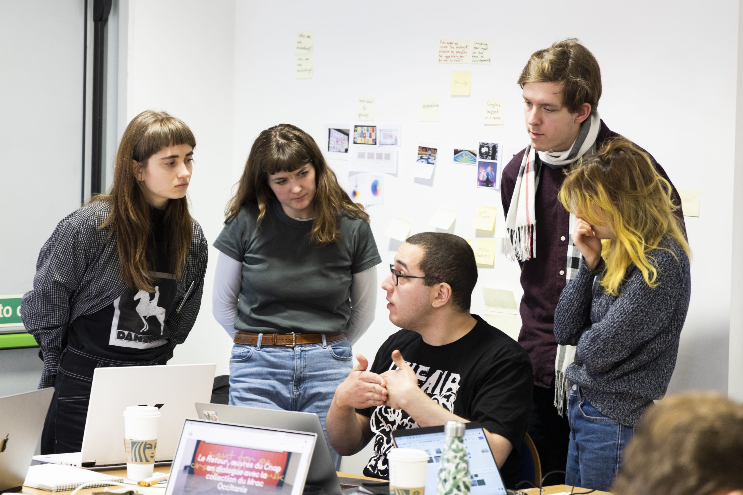  A small group of individuals engaged in a focused discussion around a table with laptops and notes. One person seated at the table is speaking, gesturing with their hands, while others standing around listen attentively. The background features sticky notes and printed materials pinned to the wall, indicating a creative or planning session.
